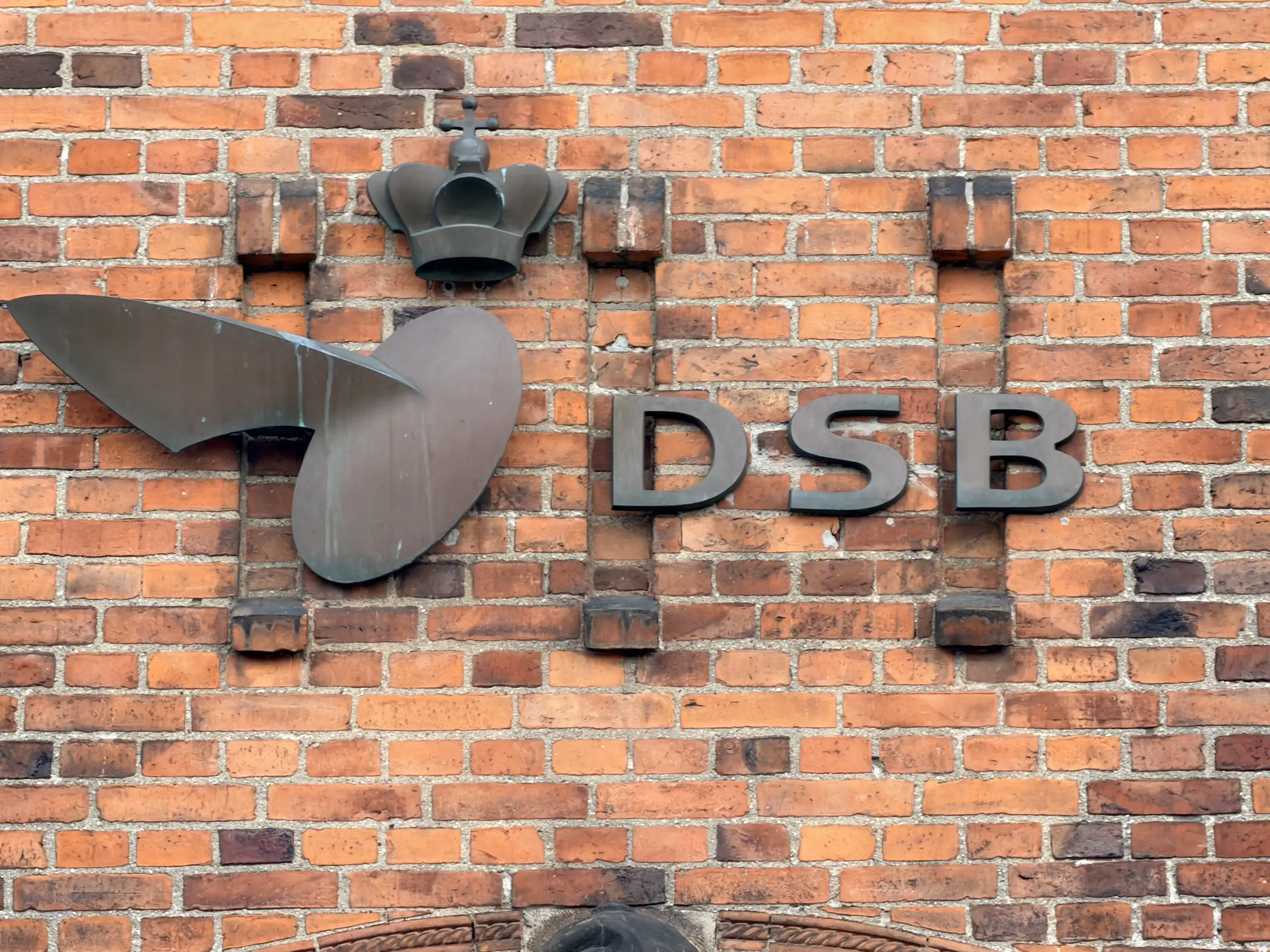 Et DSB-skilt i smedejern med vinget hjul, monteret på rød mur.