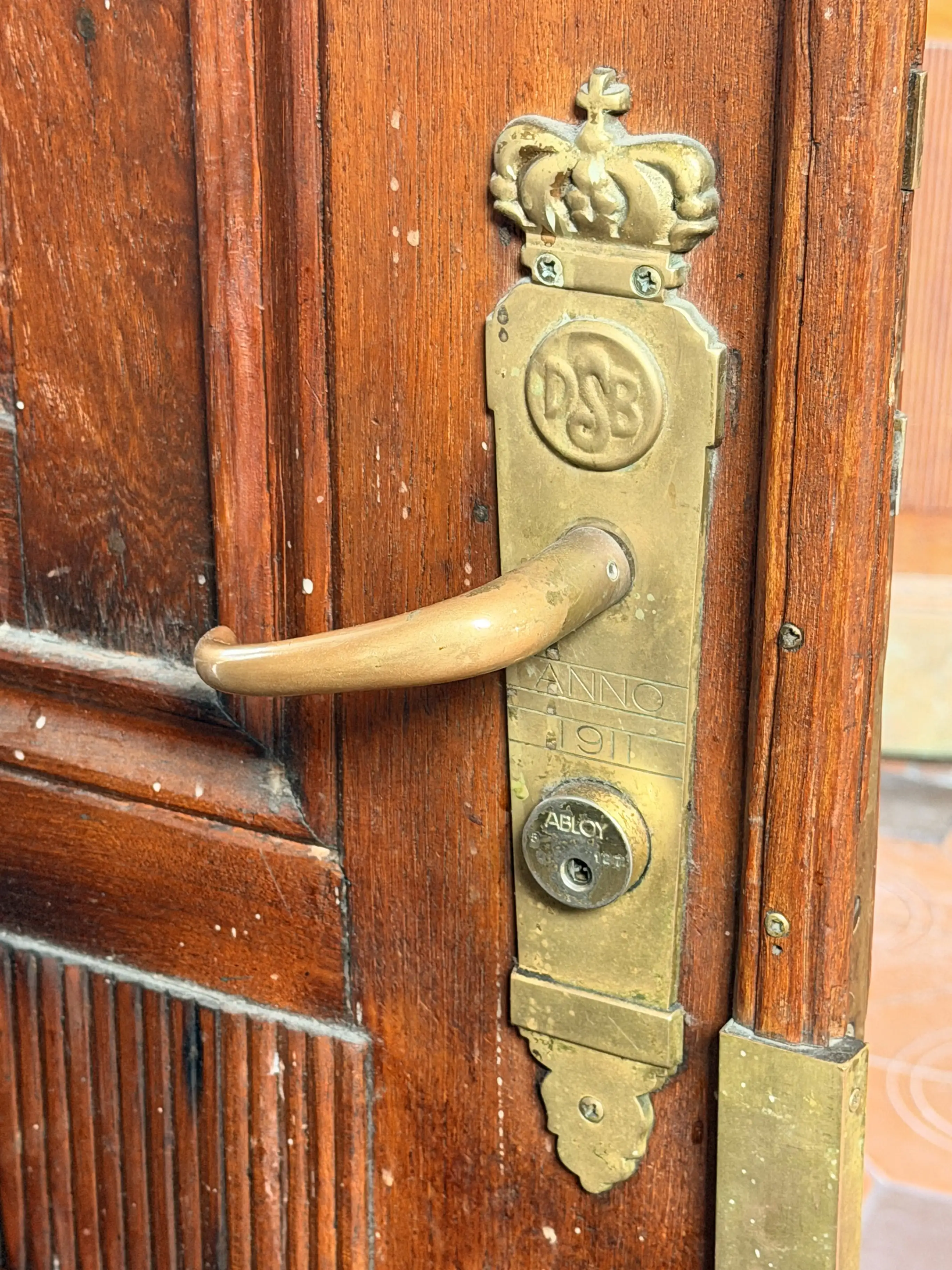 Et messinglåseskilt med DSB-monogram og årstallet 1911 på en mahognifarvet fyldingsdør.