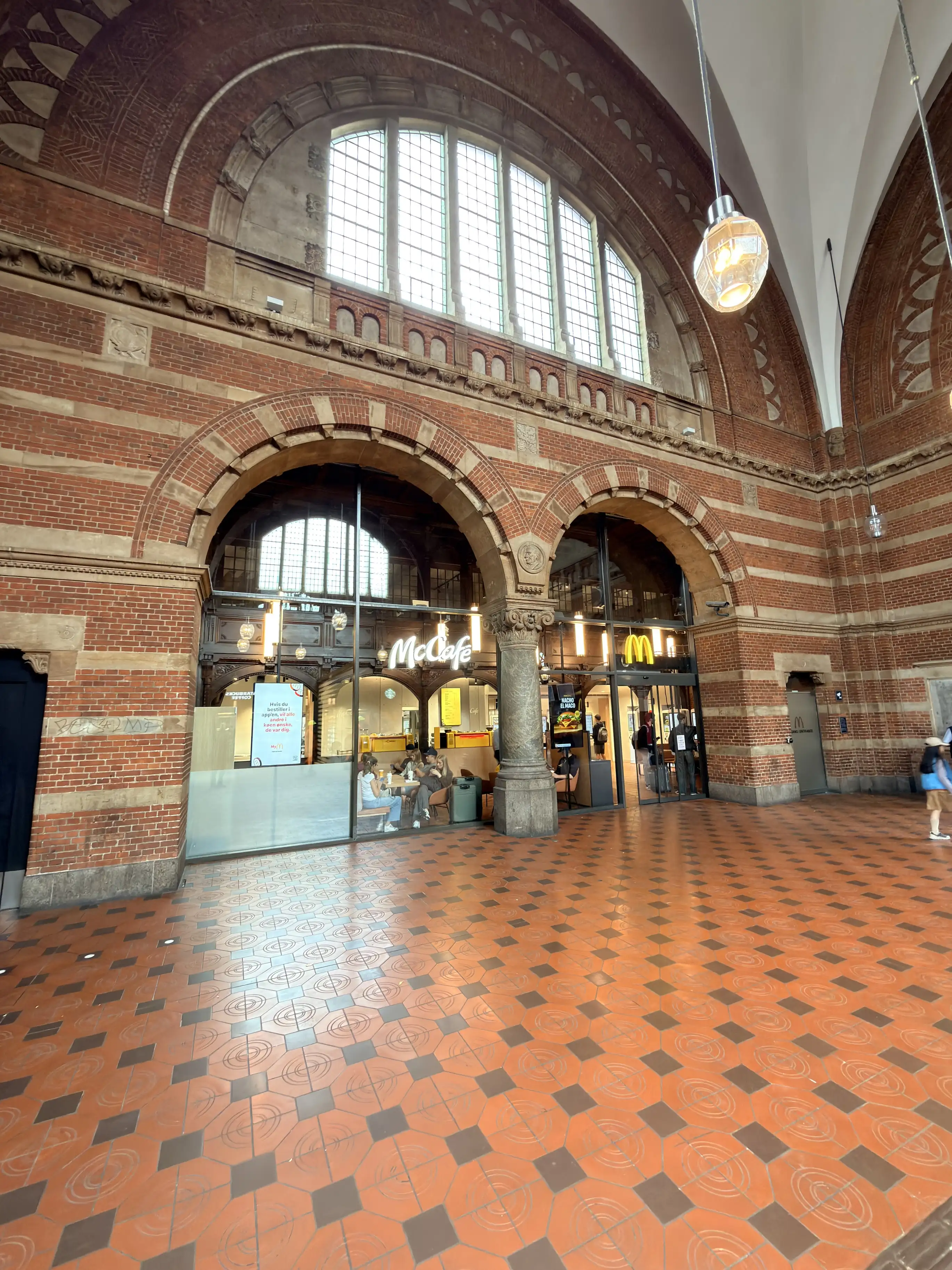 Buer i tegl og sten inde i banegården med glasfacader til butikker nedenunder og et stort halvrundt vindue ovenover.