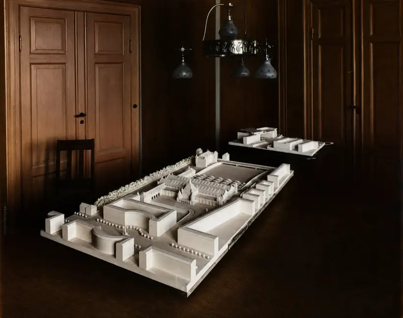 Stor hvid arkitekturmodel af et baneanlæg på et bord i et mørkt, panelklædt rum.