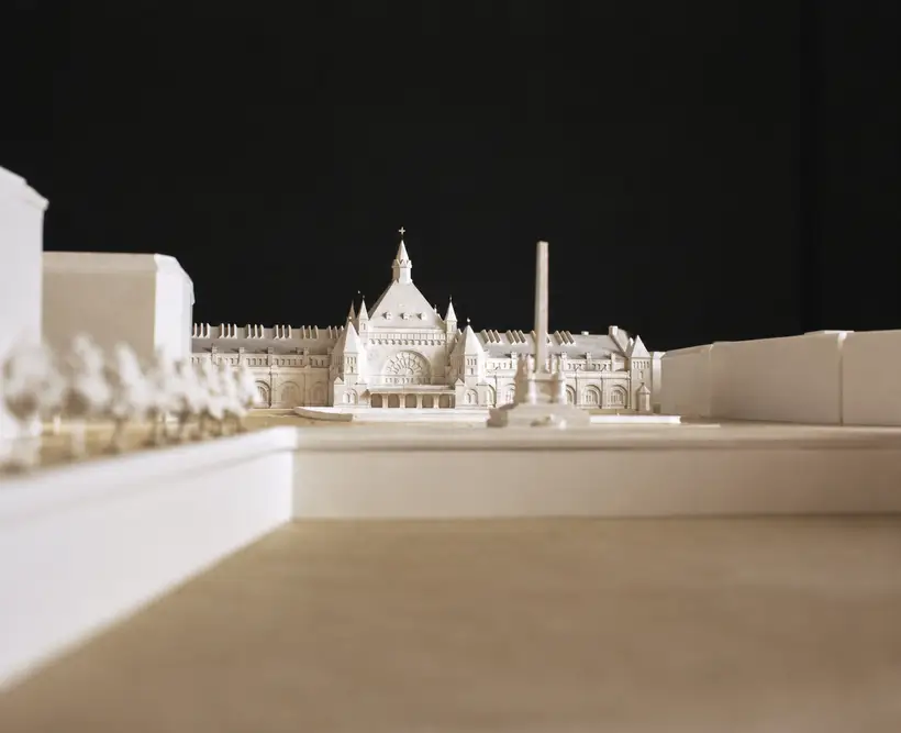 Hvid arkitekturmodel set nedefra: midterbygning med spir og rosevindue, lave sidefløje og en obelisk foran.