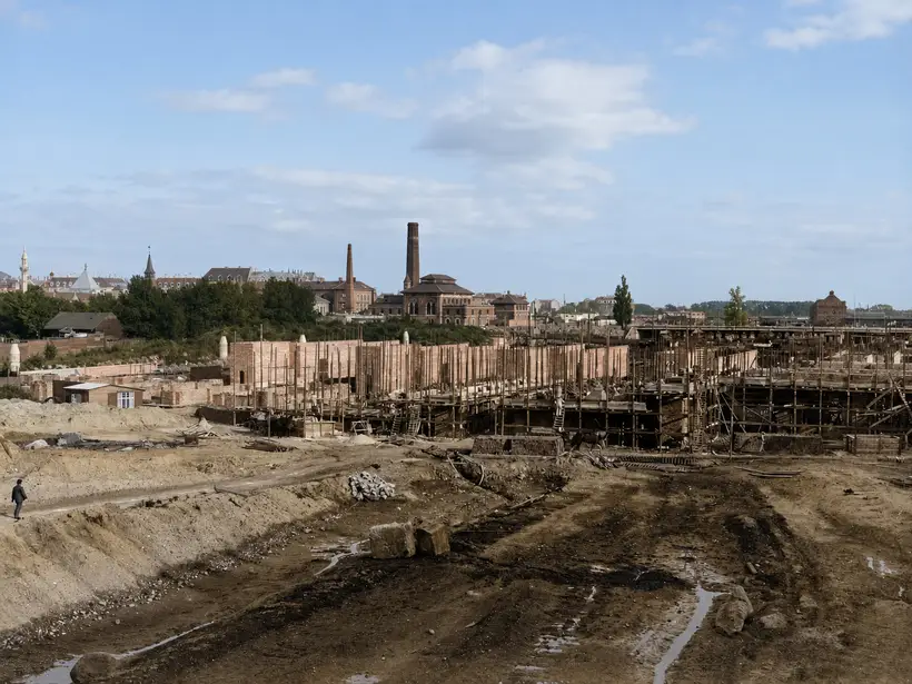 Panorama over en stor byggeplads. Lange teglmure løber tværs gennem billedet med høje stilladsstænger af rundt tømmer og arbejdsplatforme i to niveauer. Forrest opkørt mudder og vandpytter, i baggrunden fabriksskorstene og en kuppel.