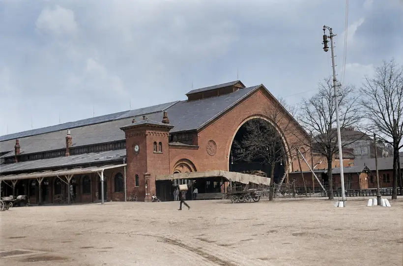 Rødstensbanegård med høj, buet gavl, urtårn og lav perronoverdækning set hen over en åben plads.