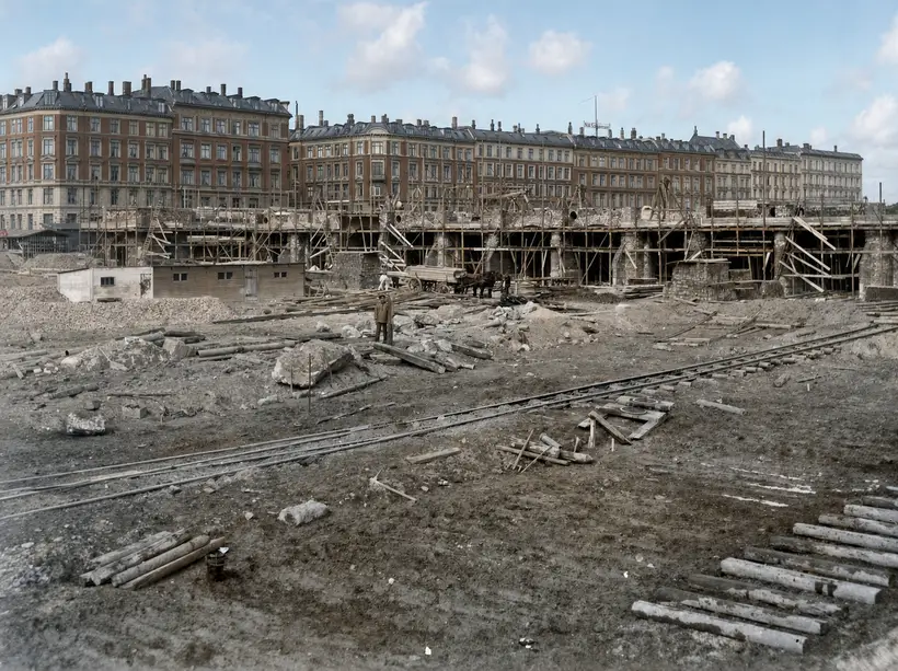 Bred byggeplads med en lang række tunge piller og buede åbninger bag et tæt rundtømmerstillads. Hestevogn med lange brædder, træskure til venstre, arbejdsspor og spredt tømmer i forgrunden.