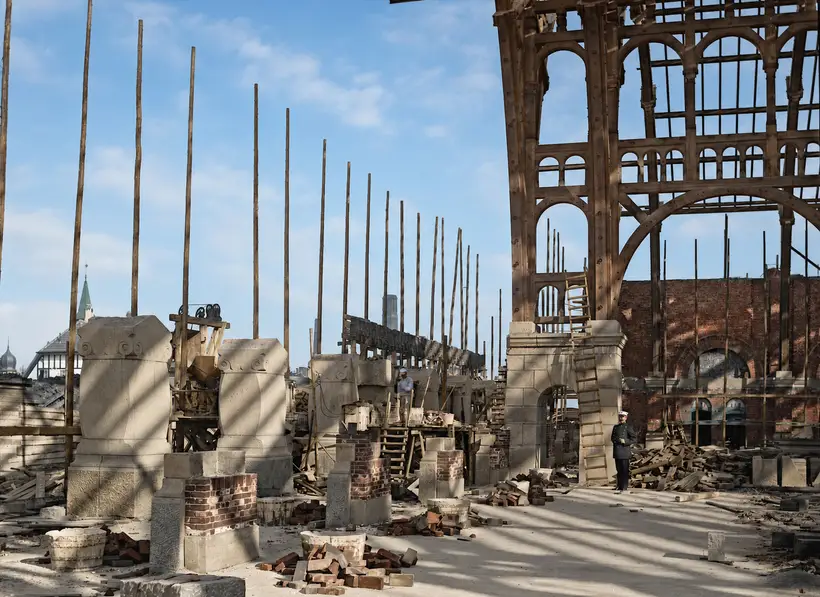 Byggeplads i højde med dækket. Stor buet trækonstruktion med gitterværk til højre, tilhugne stenblokke i forgrunden og en række høje tynde stilladsstolper.