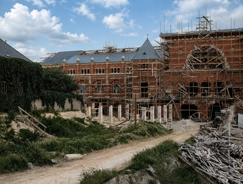 Grusvej gennem byggepladsen. I baggrunden en teglstensfløj med skifertag og åbent tagværk bagved, til højre en gavl i tæt stillads, forrest en stor stabel brædder.