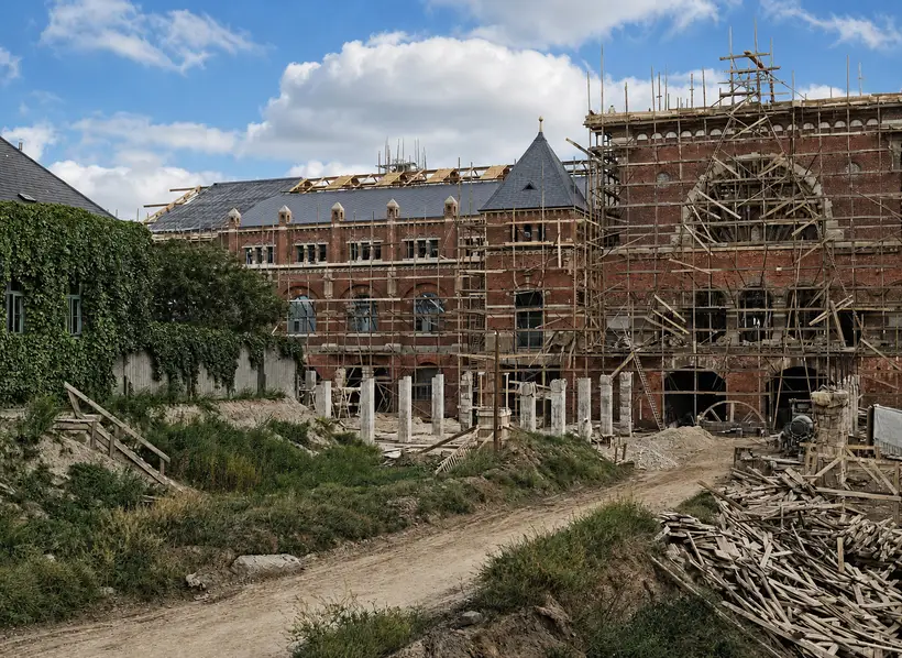 Teglstensfløj med skifertag og åbent lyst tagværk bagved; til højre en høj gavl i tæt stillads om en stor bue. Forrest grusvej og stablede brædder.