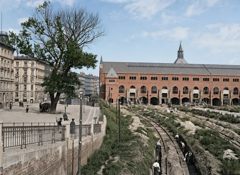 Set oppefra: til venstre en brolagt plads med gelænder, træer og høje karrébygninger; til højre en dyb udgravning hvor mænd arbejder langs et smalt spor. Bagved en lang rødstensbygning med store buer forneden og stort skifertag.