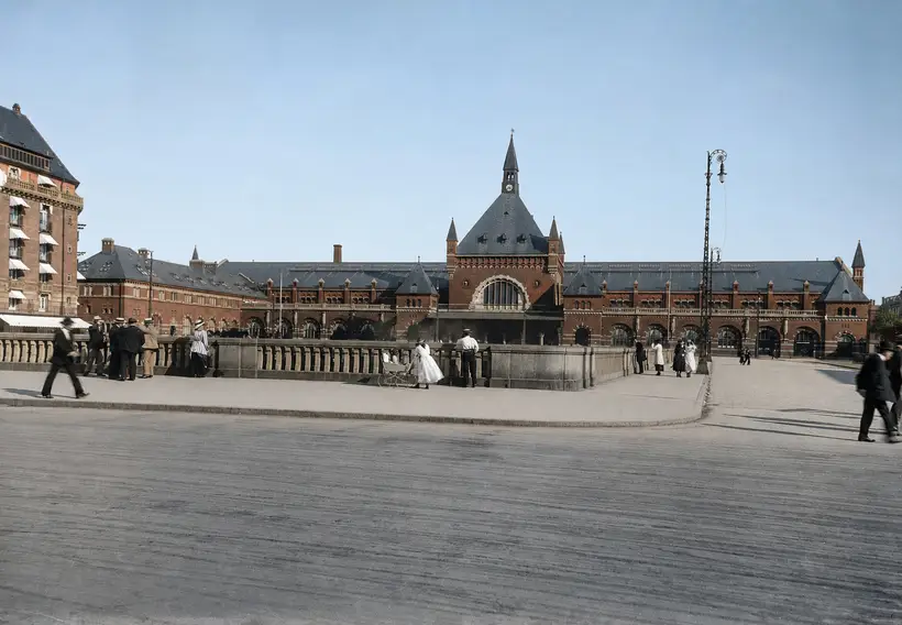 Banegårdens lange facade set over en åben plads. Midt i billedet en høj gavl med stor rundbuet rude og spir over. I forgrunden en stenbalustrade med gående mennesker i tidstypisk tøj.