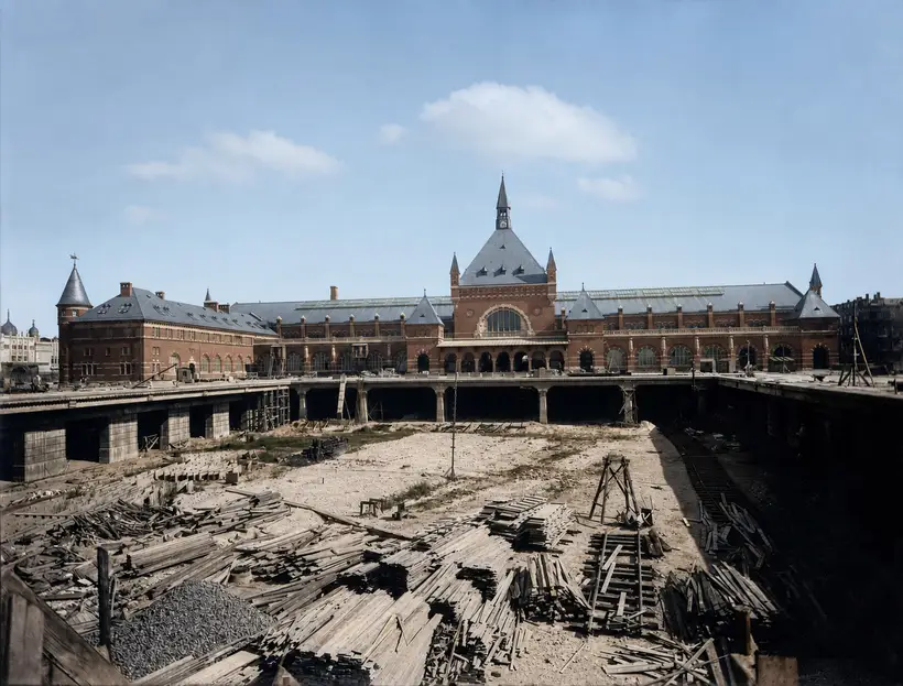 Banegårdens færdige facade set hen over en stor åben udgravning. Et dæk båret af piller løber tværs over graven. Bunden er dækket af stabler af tømmer, og en tømmerbuk står midt i den.