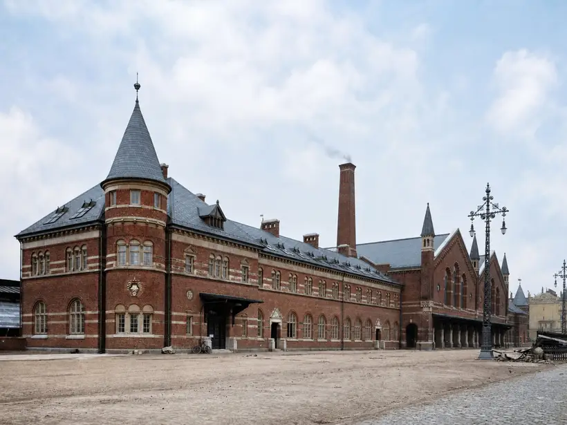 Lang rødstensfløj med rundbuede vinduer, kegleformet hjørnetårn og høj skorsten, set langs en ubefæstet vej.