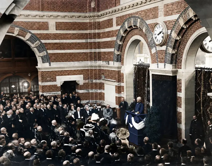 Stor forsamling i frakker og hatte omkring en taler ved en draperet talerstol under murede buer og et stort ur.