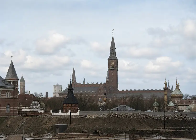 Opgravet byggeplads med lange bunker af tømmer på skråningen; bagved en mur, bare træer, rådhustårnet og løgkupler.