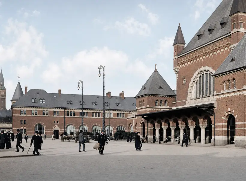Folk i lange frakker og hatte går over en brolagt forplads foran banegårdens arkade og indgangsparti.