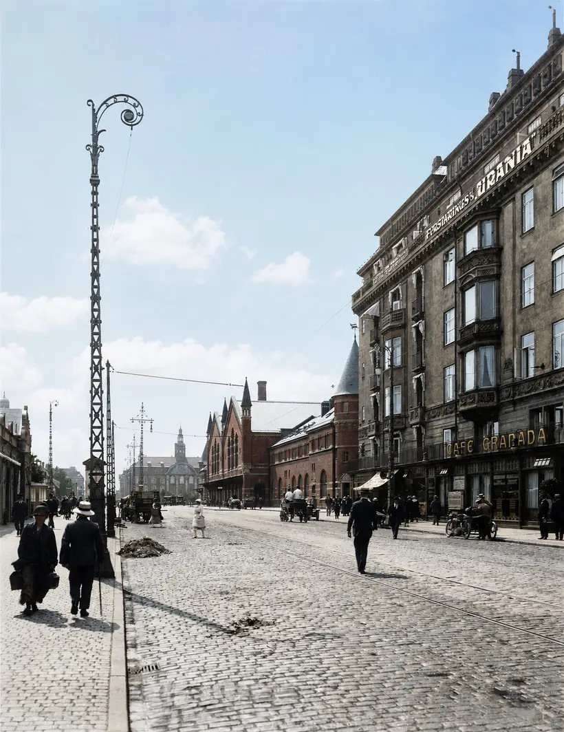 Bred brolagt gade med sporvognsskinner, høj gittermast, fodgængere og en etagebygning med butiksskilte til højre.