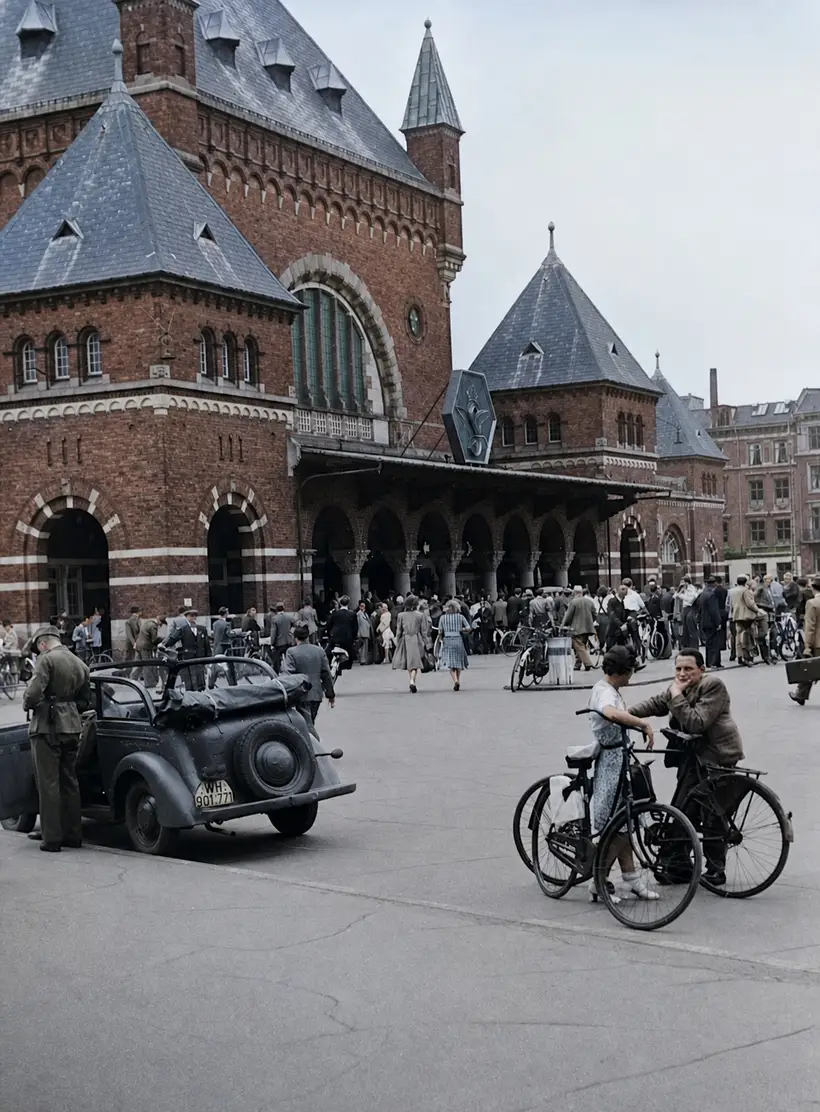 Menneskemængde og cykler foran stationens røde murstensfacade med arkade og skifertage; en åben militærvogn holder til venstre.