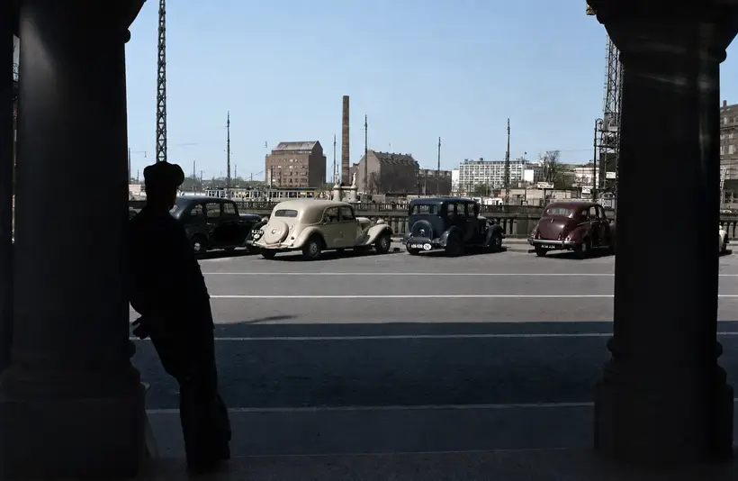 Udsigt mellem to mørke søjler mod en gade med parkerede biler fra 1940erne, en balustrade og en obelisk i baggrunden.