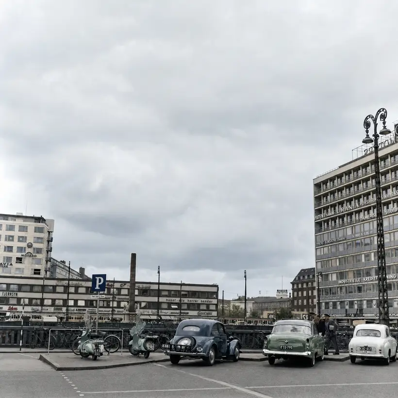 Åben plads med parkerede knallerter, cykler og biler foran høje kontorbygninger fra 1950erne under en grå himmel.