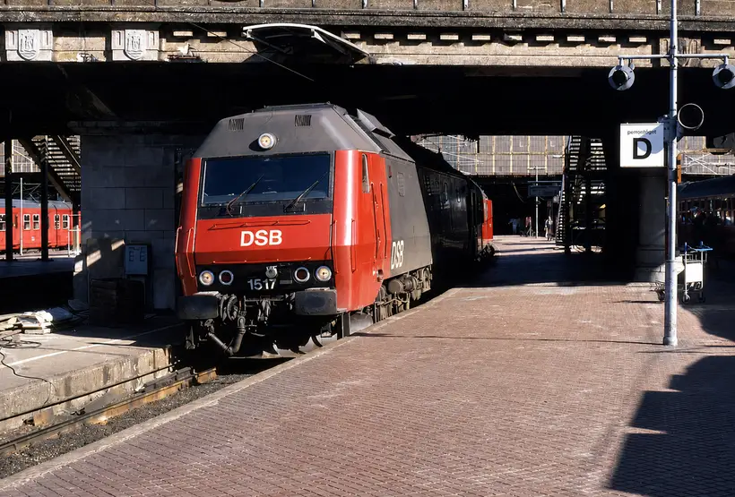 Rødt og gråt DSB-lokomotiv holder i skygge under en vejbro over sporene, med brolagt perron og trapper ned fra broen.