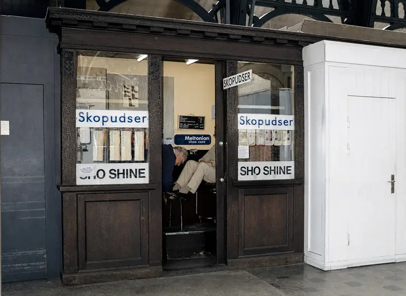 Nærbillede af udskåret trækiosk med skiltene Skopudser og SHO SHINE; indeni pudser en mand en siddende kundes sko.