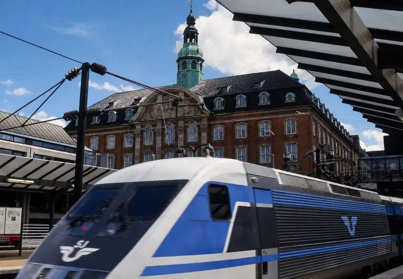 Et blåt og hvidt eltog ved en perron. Bag det en rød murstensbygning med sort tag, kviste og et grønt kobbertårn med ur.