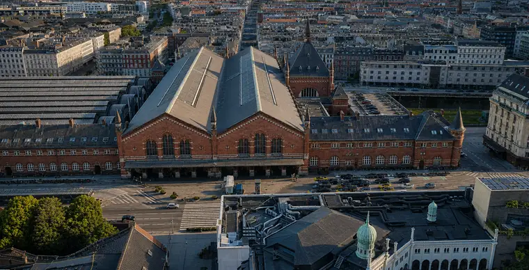 Lavt luftfoto af Hovedbanegårdens indgangsfacade mod Bernstorffsgade i varmt aftenlys: to store teglgavle med høje rundbuede vinduer og lyse sandstensbånd, en arkade bag en fremskudt baldakin i gadeplan, det tunge tårn med stejlt pyramidetag og højt spir mod nord, teglfløje til begge sider og de buede perronhaltage til venstre.