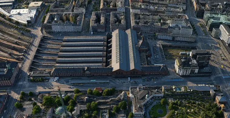 Stejlt luftfoto fra øst: seks lange buede haltage med ovenlys ligger side om side med hovedhallen for enden, røde teglfløje står mellem hallerne og Bernstorffsgade, og sporene breder sig ud i banegraven mod syd, med Tivolis haver langs billedets nederste kant.