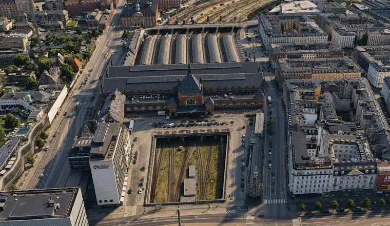 Luftfoto fra nord af Københavns Hovedbanegård: den gavlprydede indgangsbygning med spir vender mod kameraet, seks buede haltage løber væk bagved, og i forgrunden fører en åben banegrav sporene ind under gaden gennem åbninger i et betondæk, med fem- og seksetages karreer på begge sider.