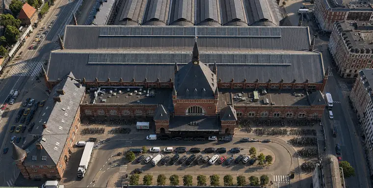 Luftfoto af Hovedbanegårdens nordfacade: rød tegl med sandstensdetaljer, en kamtakket midtergavl med ur og slankt spir, lange fløje til begge sider, hovedhallens tag bagved og de buede perronhaller længst væk. Biler, træer og lange rækker af cykler fylder pladsen foran.