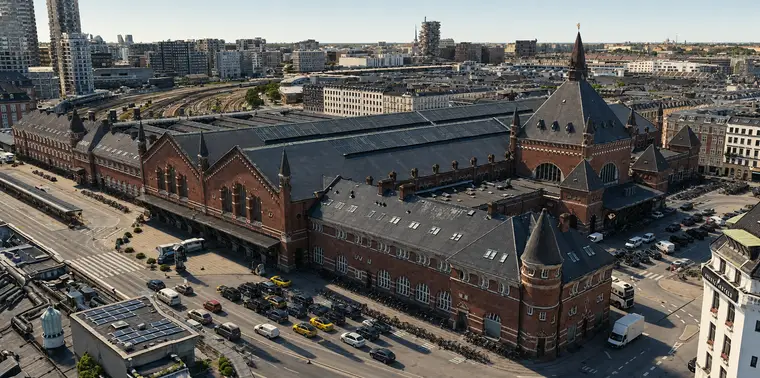 Københavns Hovedbanegård fra nordøst, lavt over tagene. En lang rødstensfløj med rundbuede vinduer følger Bernstorffsgade, og nedenfor holder taxaer og lange rækker af cykler. Til højre rejser indgangstårnet med sit sorte spir sig over hovedfacadens store buevindue. Bagved ligger perronhallernes glastage, og i baggrunden breder sporene sig ud i en vifte.