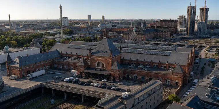 Københavns Hovedbanegård fra nordvest under en klar himmel. Hovedfacaden står midt i billedet med en murstensgavl, et stort buet vindue og et slankt tårn med spir. Foran ligger en forplads med parkerede biler og en lastbil. Nedenfor løber flere spor i en åben grav under en vejbro på betonpiller. Til højre er de lange haltage dækket af solceller. Til venstre ses Tivolis forlystelser og trætoppe, og i horisonten står byggekraner over byen.