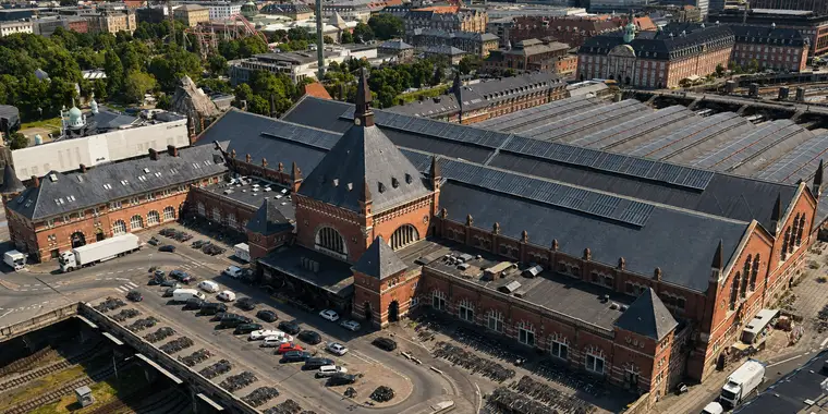Københavns Hovedbanegård fra nordvest, tæt på og stejlt oppefra. Murstensfacaden fylder den nederste halvdel med et højt tårn med spir, et stort buet vindue og en hjørnepavillon. Bagved bærer et bredt skifertag tre lange striber solceller, og videre bagude løber seks buede glastage mod højre. På forpladsen står biler og lange rækker af cykler. Øverst til venstre ligger Tivolis træer, grønne kupler og en rutsjebane.