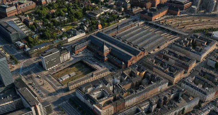 Højt luftfoto fra nordvest: hovedhallen krydser billedets midte med den spirprydede indgangsbygning på nordsiden, de seks perronhaller bagved og sporene, der breder sig ud forbi dem. En banegrav løber mod kameraet, Tivoli ligger øverst til venstre, og tætte 1800-talskarreer fylder den nederste halvdel.