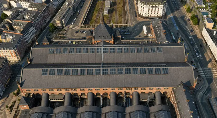 Næsten lodret luftfoto af hovedhallens tag: en bred grå flade med to lange ovenlysbånd og små spirprydede murtårne i hjørnerne, nordindgangens stejle tage og spir bagved, og langs nederste billedkant de seks hvælvs runde ender mod en høj mur gennembrudt af rundbuede åbninger.
