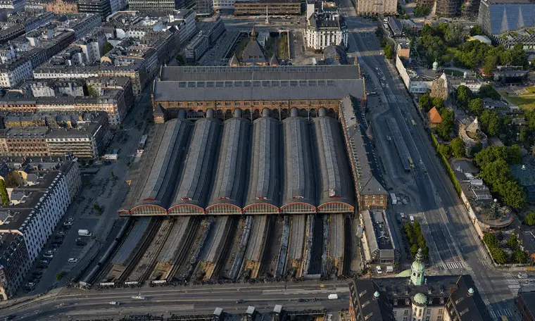Luftfoto fra syd: seks halvcirkelformede glasgavle med rødmalede buer vender mod kameraet, de seks tøndehvælv med ovenlysbånd løber mod nord bag dem til hovedhallens brede tag, og foran ligger sporene sænket mellem perronerne og løber ud mod gaden.