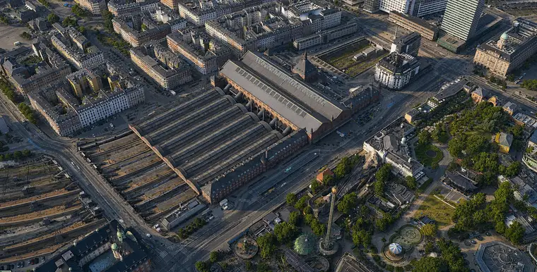 Højt luftfoto fra sydøst: de seks perronhaller med deres buede gavle ligger nederst til venstre, sporene breder sig ud i en åben banegrav bagved, hovedhallen krydser for enden med det spirprydede tårn ved siden, og en anden banegrav fører banen videre mod nord mellem husene, med Bernstorffsgade langs facaden og Tivoli overfor.