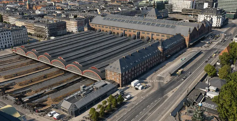 Lavt luftfoto af de seks perronhallers sydender: seks buede glasgavle på række, hver bue indrammet af et bredt rødt bånd over en lys sålbænk med en lille rød rygningsventil bag toppen, de lange mørke haltage løbende væk bagved og hovedhallens tag og nordtårnets spir længst væk.