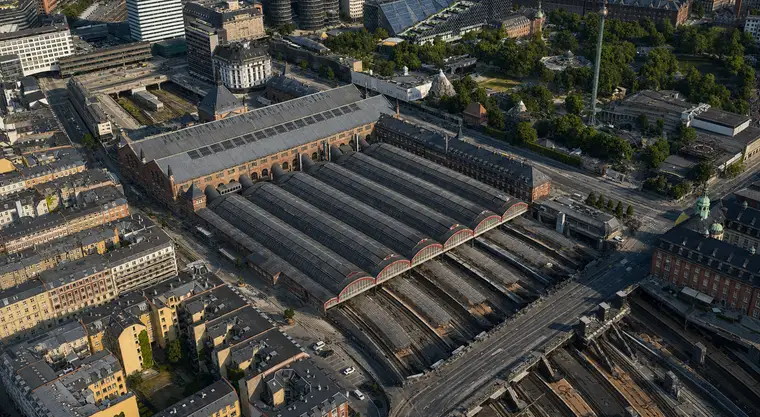 Luftfoto fra sydvest: hovedhallens lange grå tag ligger øverst til venstre med ovenlysbånd og hjørnetårne, de seks tøndehvælvede perronhaller med ovenlysstriber løber ned mod højre og ender i halvcirkelformede glasgavle med røde buer, og bag dem falder sporene ned i en banegrav under en vejbro. Vesterbro fylder nederst til venstre, Tivoli øverst til højre.