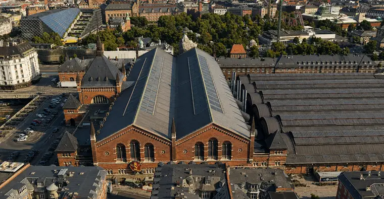 Lavt luftfoto fra vest: hovedhallens solbelyste dobbeltgavl i rød tegl står tæt på kameraet med høje rundbuede vinduer og konsolbårne gesimser, det dobbeltrygggede skifertag løber tilbage bagved, indgangsbygningen med spiret vender bort til venstre, og perronhallerne trapper af mod højre.
